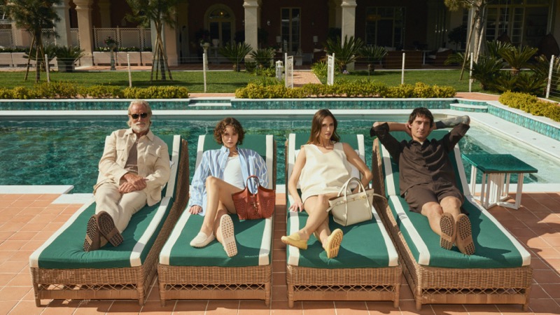 Models in Tod's summer 2026 recline on green and white striped sun loungers beside a villa pool, carrying structured leather bags and wearing Gomminos