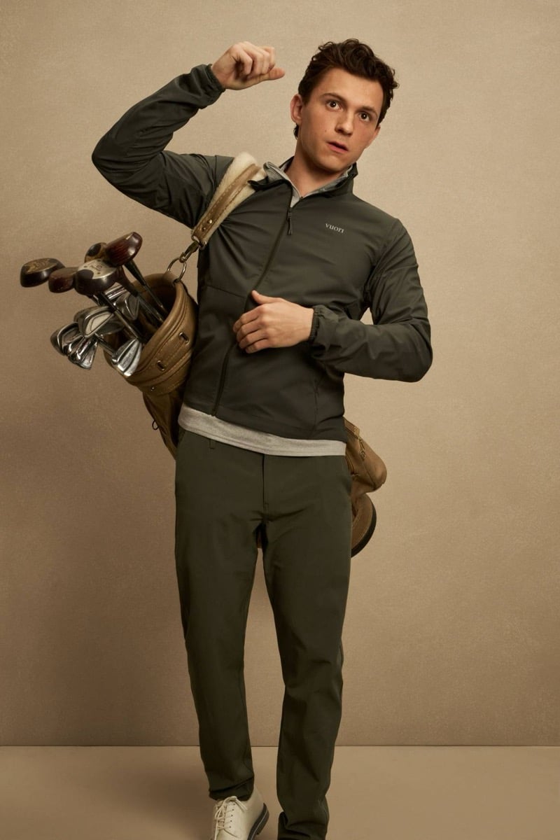 Holland wears an olive zip jacket over a gray tee with olive technical pants and a leather golf bag