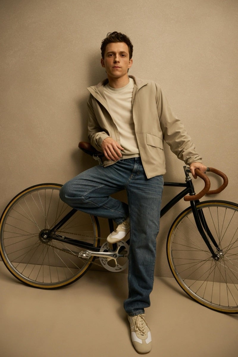 Holland leans against a wall beside a bicycle in a tan hooded jacket over a cream tee with jeans