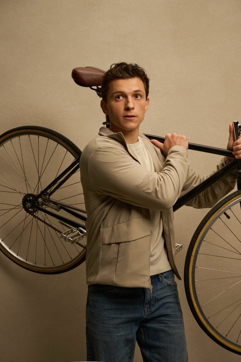Holland carries a bicycle over his shoulder in a tan hooded jacket and jeans