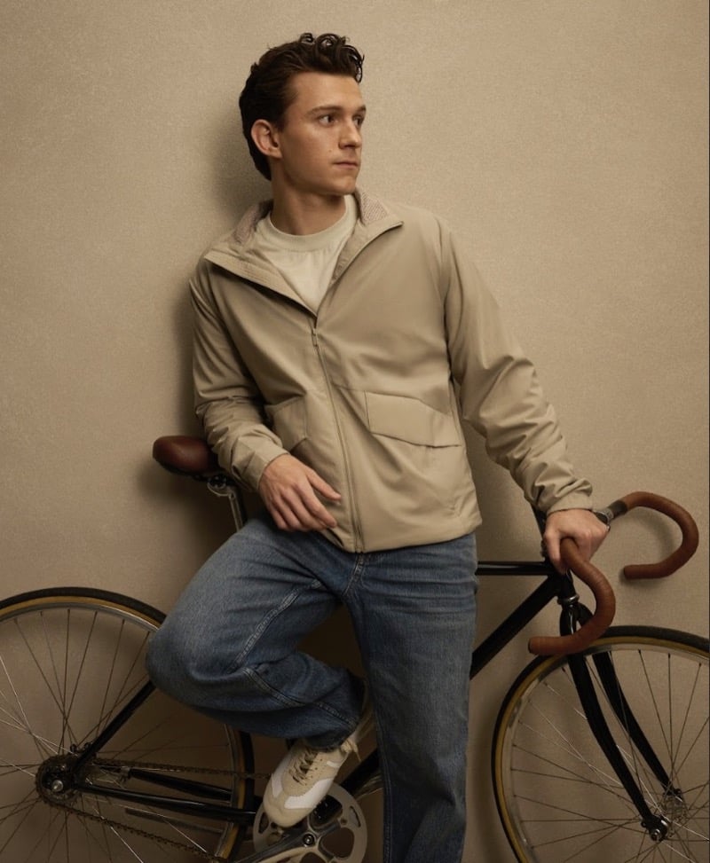 Holland leans against a wall beside a bicycle in a tan hooded jacket over a cream tee with jeans