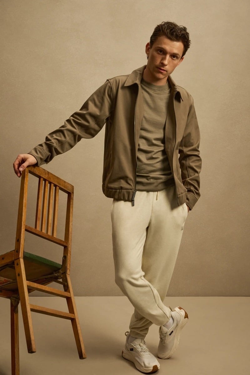 Holland leans on a wooden chair in an olive zip jacket over an olive tee with joggers