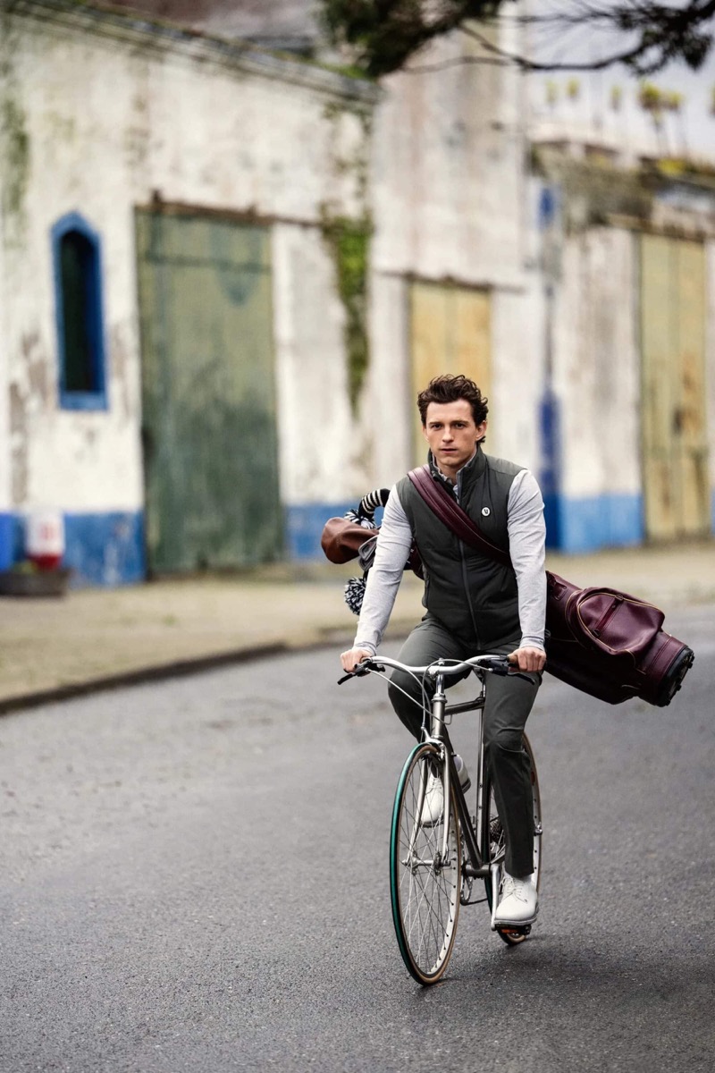 Holland rides a bicycle through Comporta in a quilted vest over a gray long-sleeve top with a leather golf bag