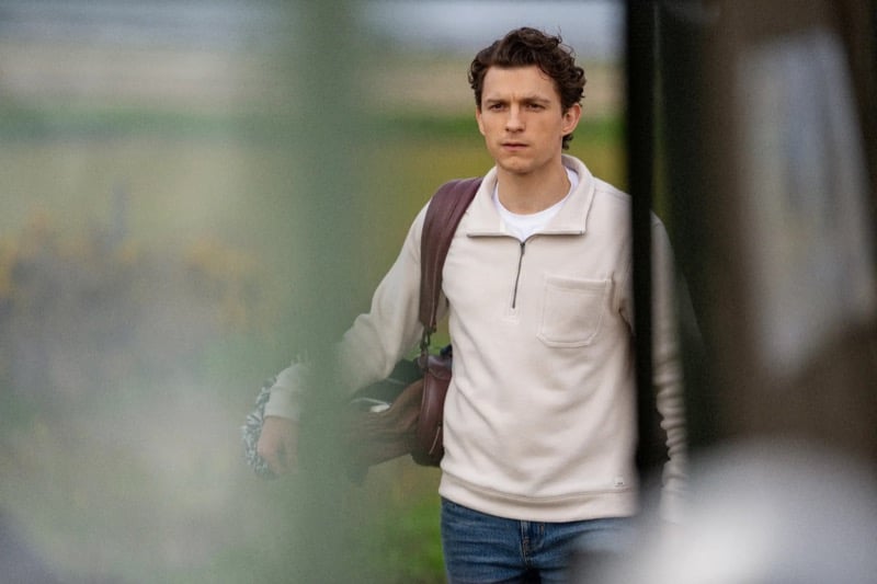 Holland wears a cream half-zip fleece over a white tee with jeans and a leather bag over his shoulder