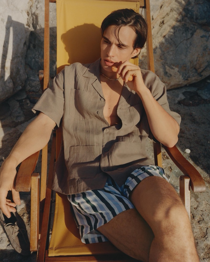 Louis wears a brown guayabera shirt over blue-striped swim shorts in a deck chair