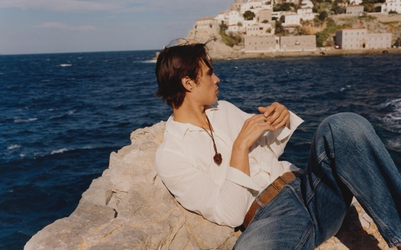 Louis reclines on a rocky shoreline in a white linen shirt and jeans with Hydra in the background