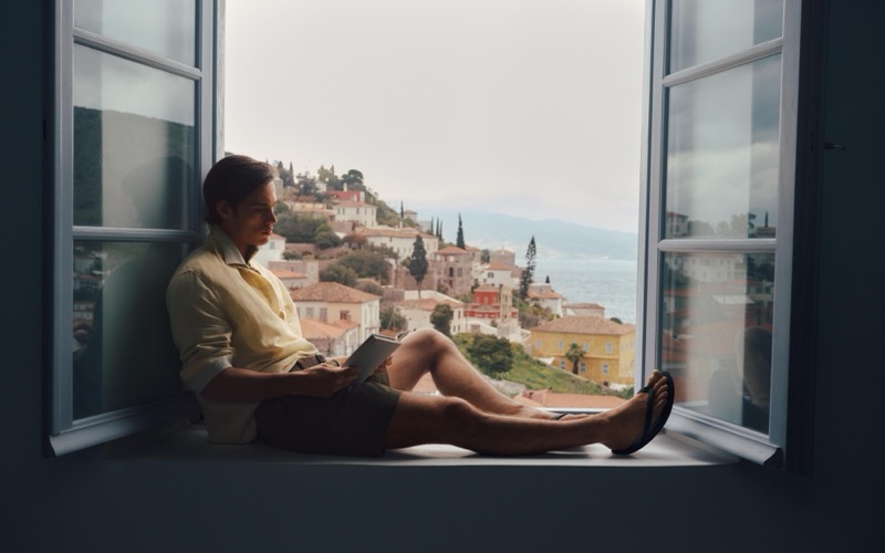 Louis reads a book in a window overlooking Hydra in a pale yellow poplin shirt and olive cargo shorts