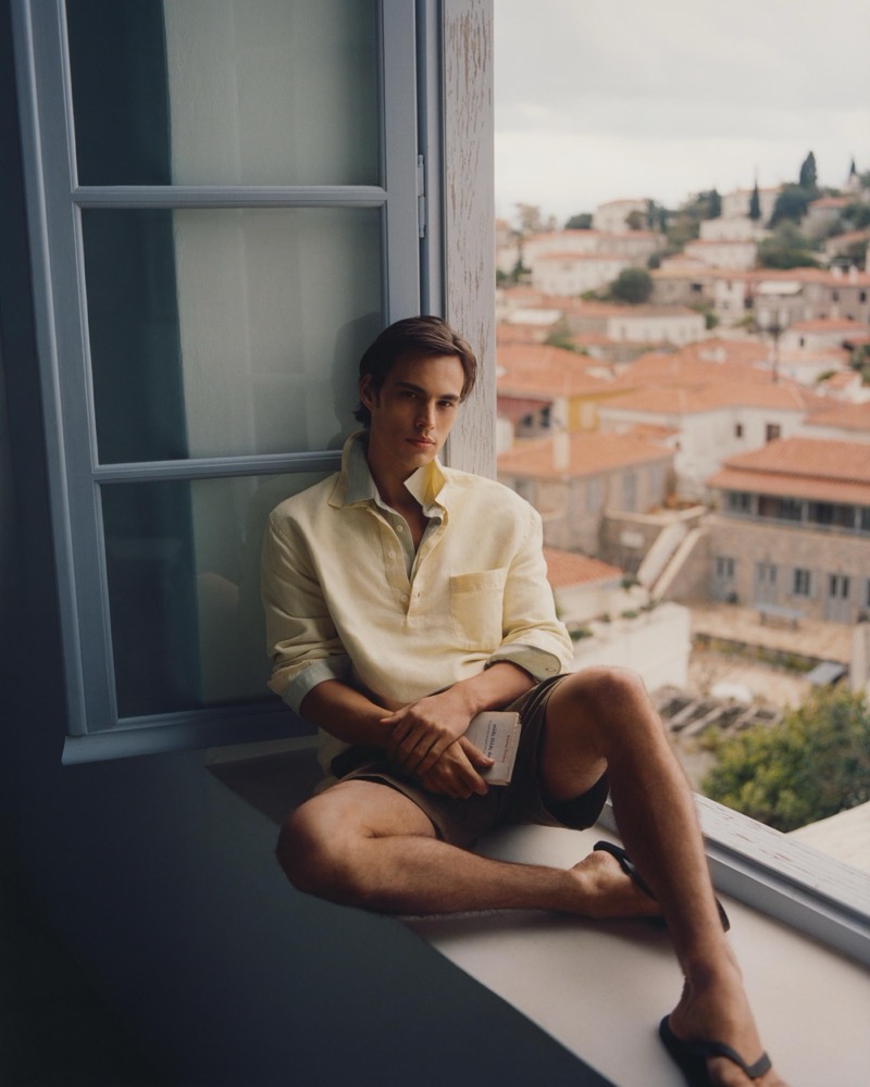 Louis sits in a window sill in a pale yellow linen shirt and dark shorts overlooking Hydra