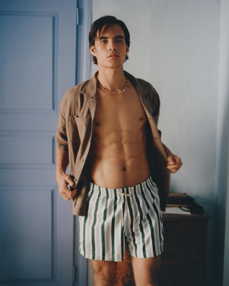 Louis wears an open sand linen shirt over green-and-white striped swim trunks with a stone pendant necklace