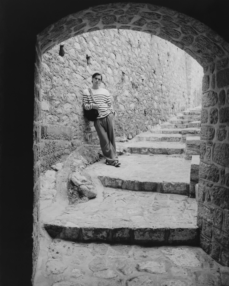 Louis stands in a stone archway wearing a Breton-stripe knit with relaxed trousers and sandals