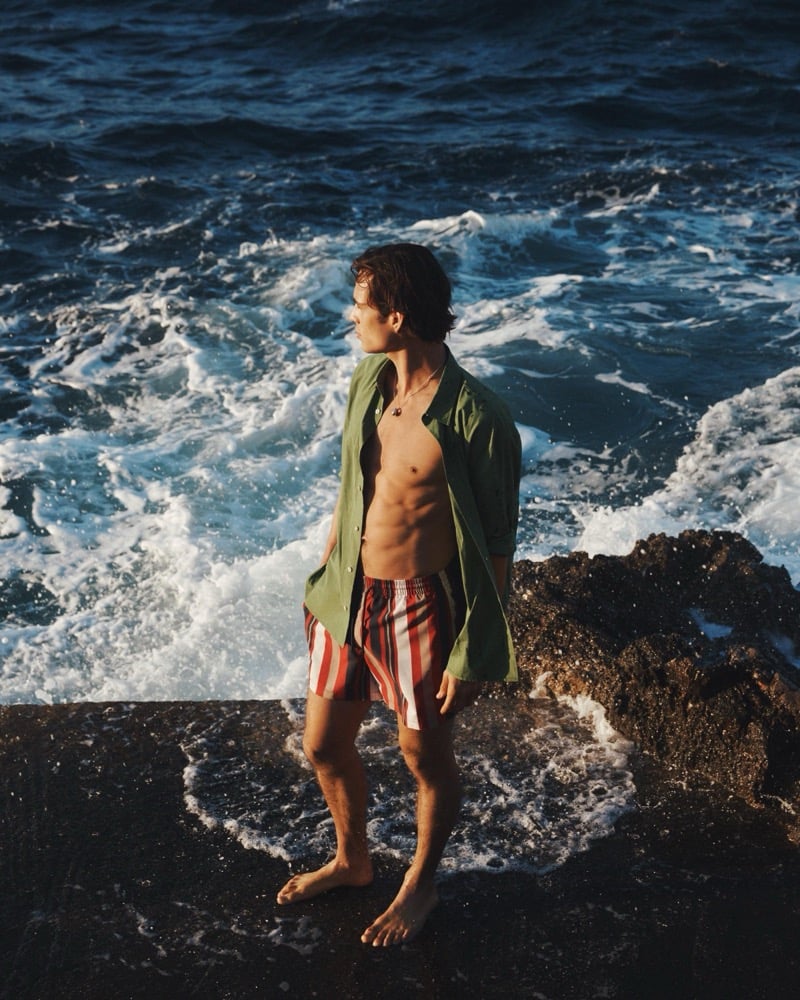 Louis wears an open olive shirt over red-striped swim shorts on the rocky shoreline
