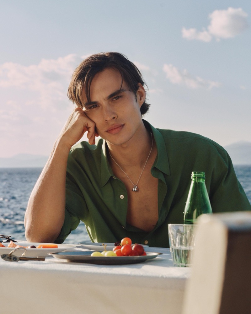 Louis wears a deep green shirt with a stone pendant necklace seated at a seaside table