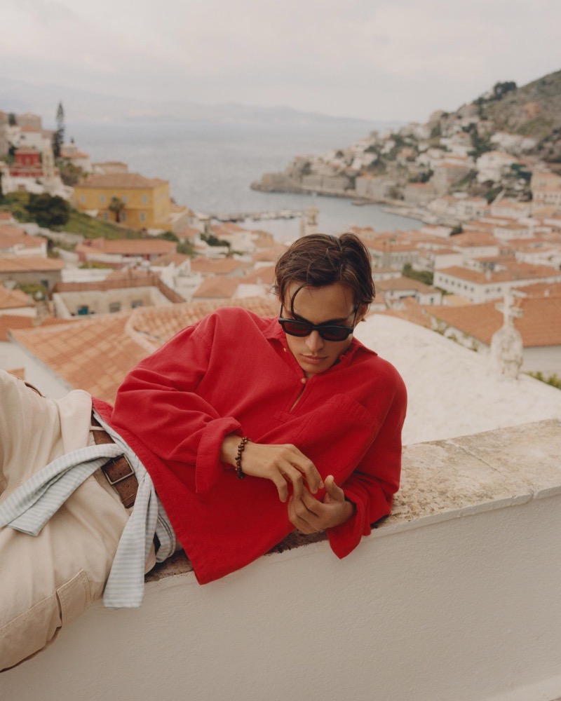 Louis wears a red linen pullover with sunglasses leaning on a terrace wall overlooking Hydra