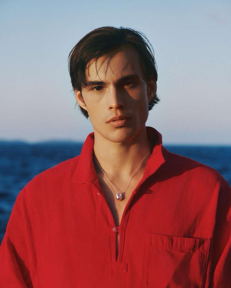 Louis wears a red linen pullover with a stone pendant necklace against the sea