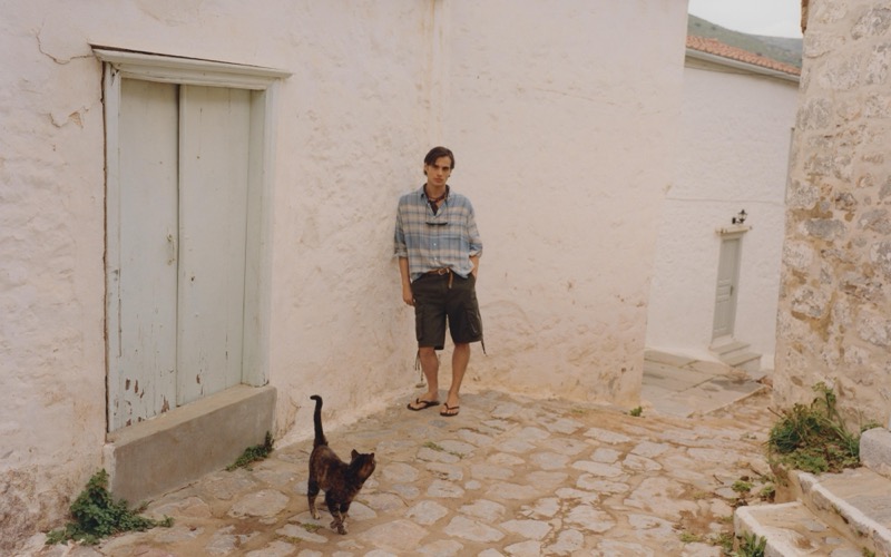 Louis wears a blue-and-sand plaid shirt with dark cargo shorts and sandals in a whitewashed alleyway