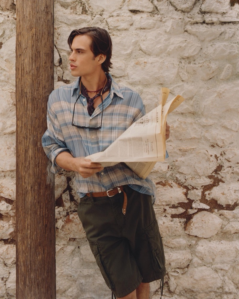 Louis wears a blue-and-sand plaid shirt open with a bandana and olive cargo shorts holding a newspaper