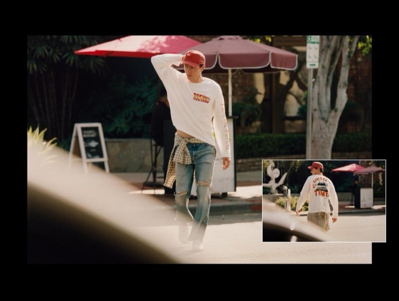 Clement walks a Los Angeles sidewalk in a white long-sleeve Back to the Future graphic T-shirt with ripped straight-leg jeans, a plaid shirt tied at the waist, and a red baseball cap