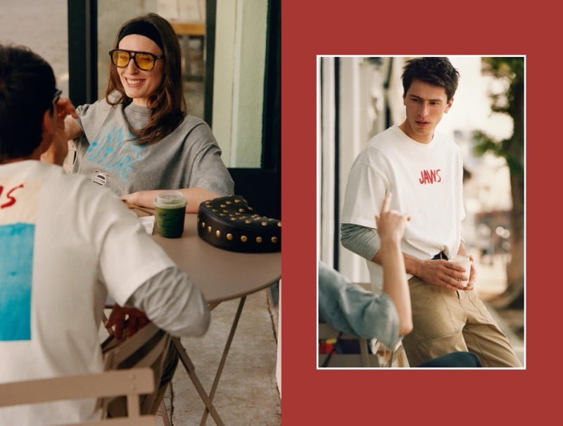 Ansley wears a grey graphic sweatshirt with yellow-lens aviator sunglasses and a black headband at a cafe, while Clement wears a white Jaws graphic T-shirt over a grey long-sleeve with khaki trousers, holding a coffee