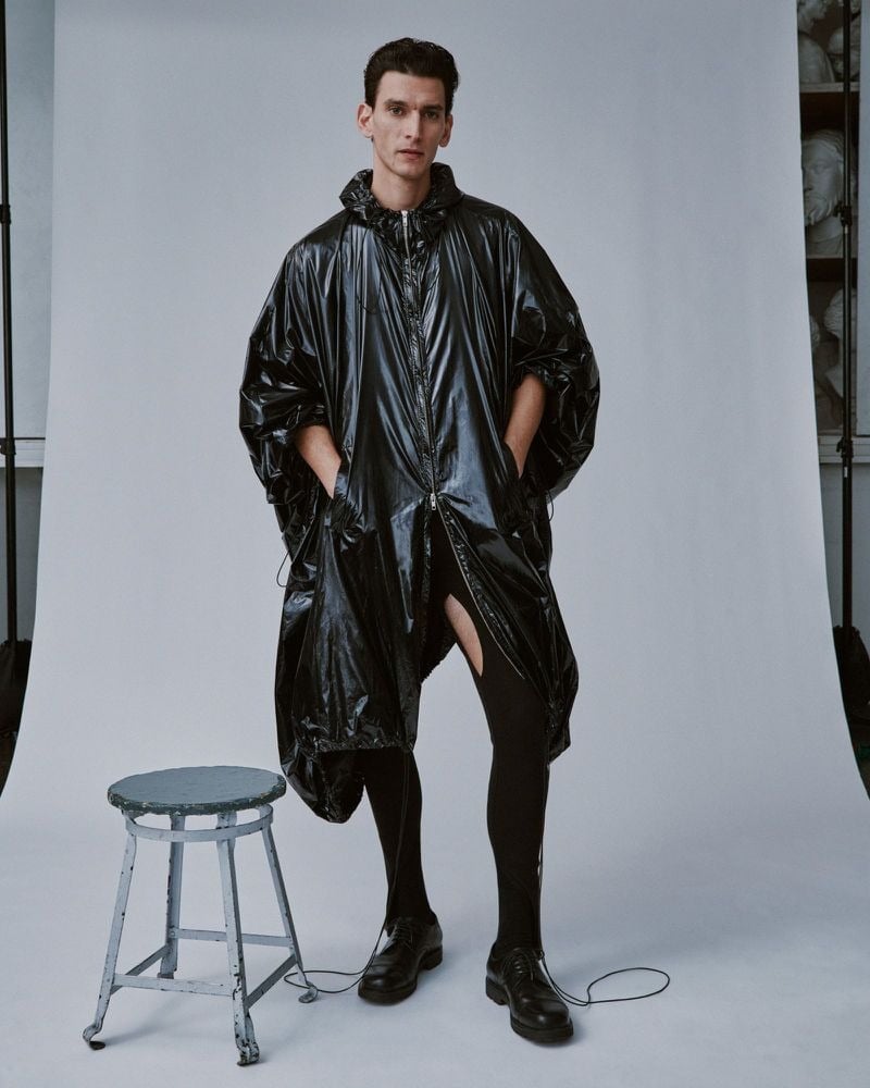 Thibaud Charon stands beside a metal stool in an L.I.I. heavy black hooded poncho in glossy coated nylon zipped to the chest with a vented split at the knee, paired with black footed leggings and black leather boots, photo studio backdrop
