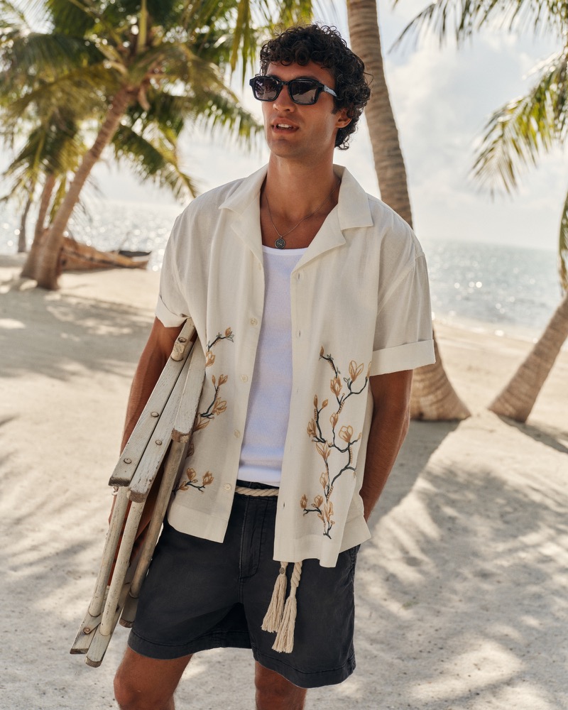 Francisco wears an open white Abercrombie & Fitch resort camp-collar shirt with botanical magnolia embroidery over a white ribbed tank and washed black pull-on shorts, holding folded wooden beach chairs on a palm-lined shore