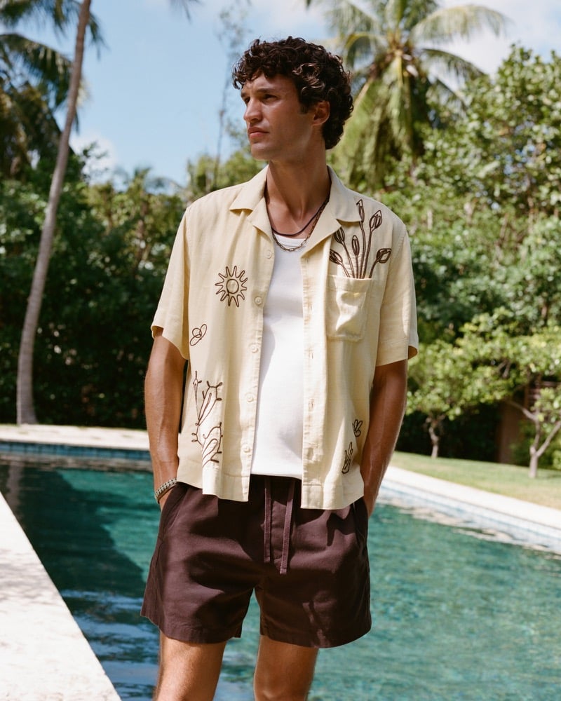 Francisco wears an open pale yellow Abercrombie & Fitch resort camp shirt with sketch-like sun and floral embroidery over a white ribbed tank and dark brown pull-on shorts, standing poolside in front of dense tropical greenery