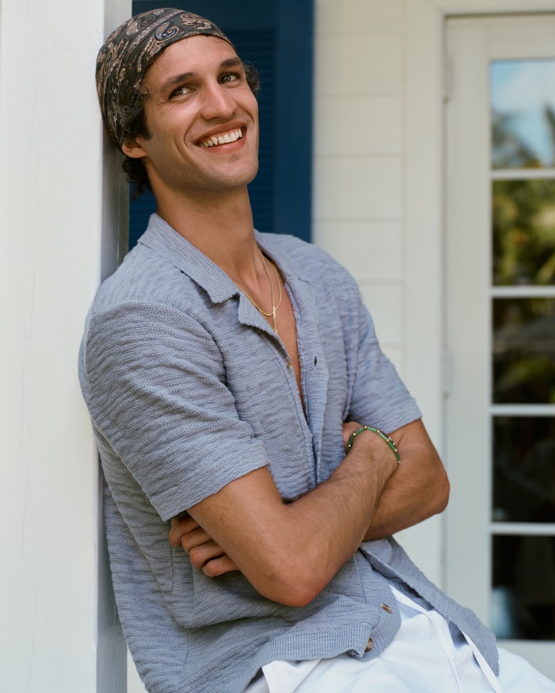 Francisco smiles in a light blue Abercrombie & Fitch cotton-knit button-through polo worn open over a bare chest with a paisley bandana tied around his head, leaning against a white-paneled wall beside a blue shuttered door