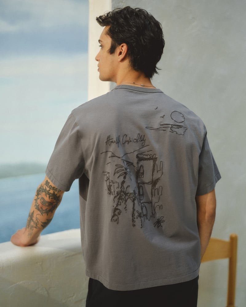 Back view of Mase in a slate grey Abercrombie & Fitch boxy graphic tee printed with an Amalfi Capri Italy line drawing of coastal architecture and a setting sun, paired with black trousers