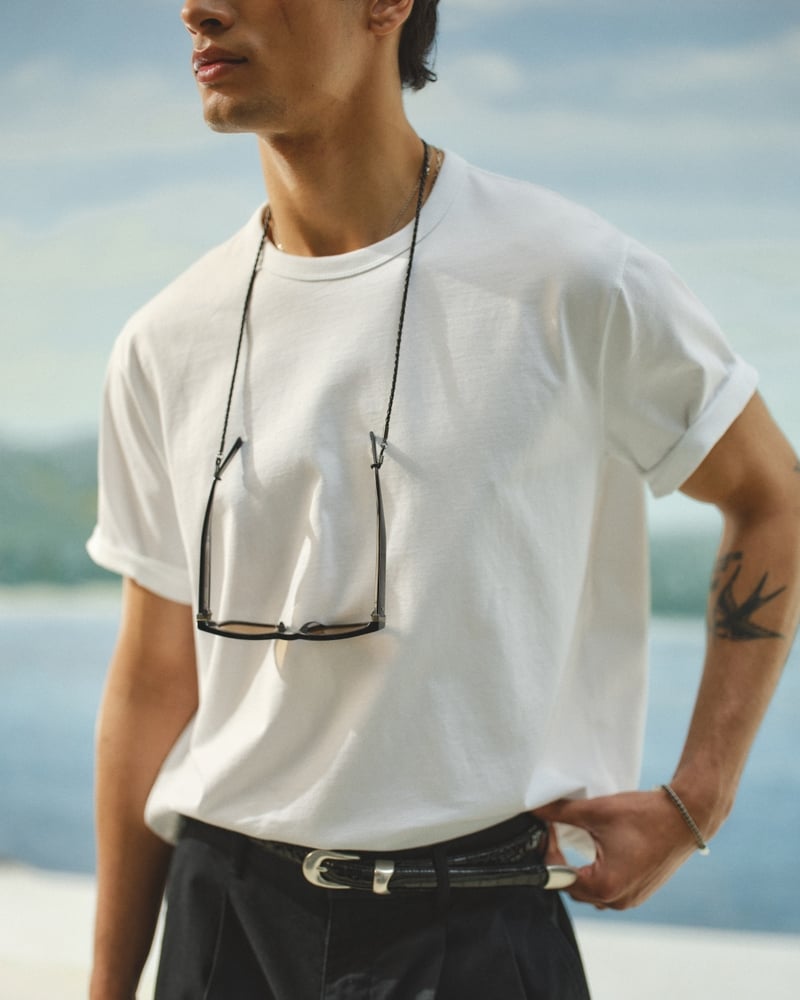 Close-up of Mase in a white Abercrombie & Fitch boxy tee with cuffed sleeves, black-framed sunglasses hanging from a cord around his neck, paired with black pleated trousers and a black leather belt