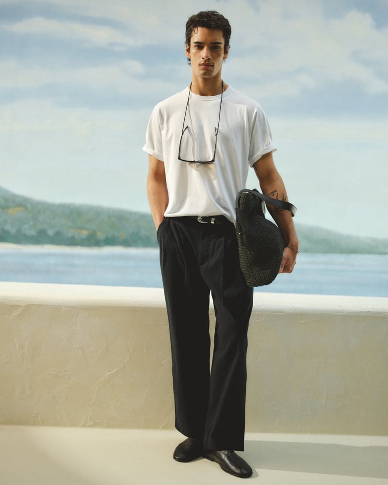Mase stands against a painted coastal mural in a white boxy tee tucked into black Abercrombie & Fitch pleated baggy trousers, holding a black woven raffia tote, with sunglasses suspended from a cord around his neck