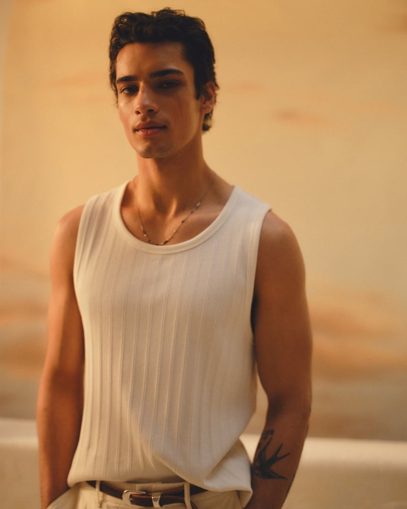 Mase stands against a warm peach-toned wall in a white Abercrombie & Fitch premium ribbed tank, cream trousers, and a brown leather belt, hands tucked into his pockets