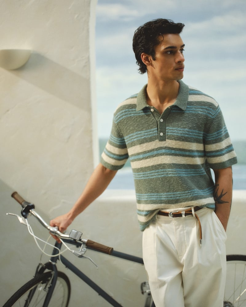 Mase stands beside a bicycle in an Abercrombie & Fitch striped knit sweater polo in sage, teal, and cream, paired with white pleated baggy trousers, a leather belt, and a tan drawstring detail