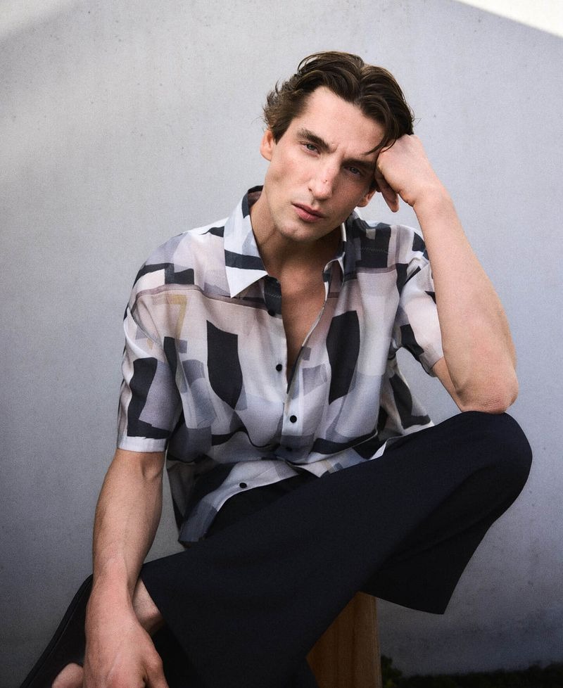 Anatol seated against a pale wall in an Adolfo Dominguez abstract-print short-sleeve shirt in gray, ivory, and black with dark wide-leg trousers