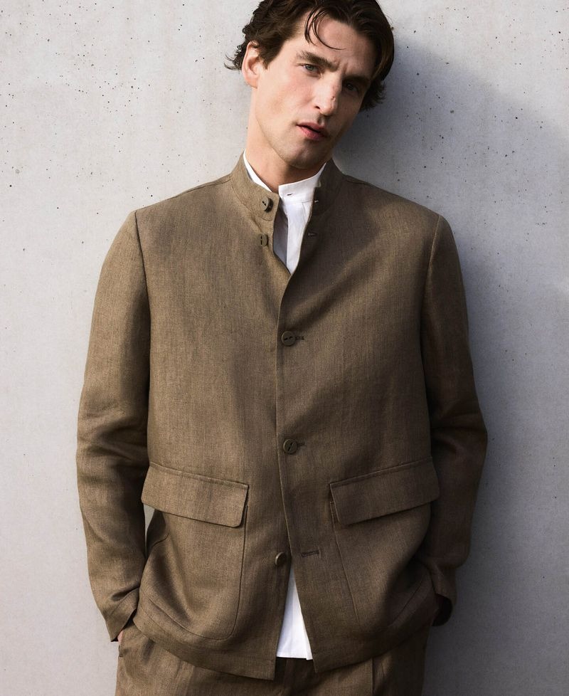 Anatol leans against a concrete wall in an Adolfo Dominguez Mandarin-collar taupe linen jacket over a white Mandarin-collar shirt with matching trousers