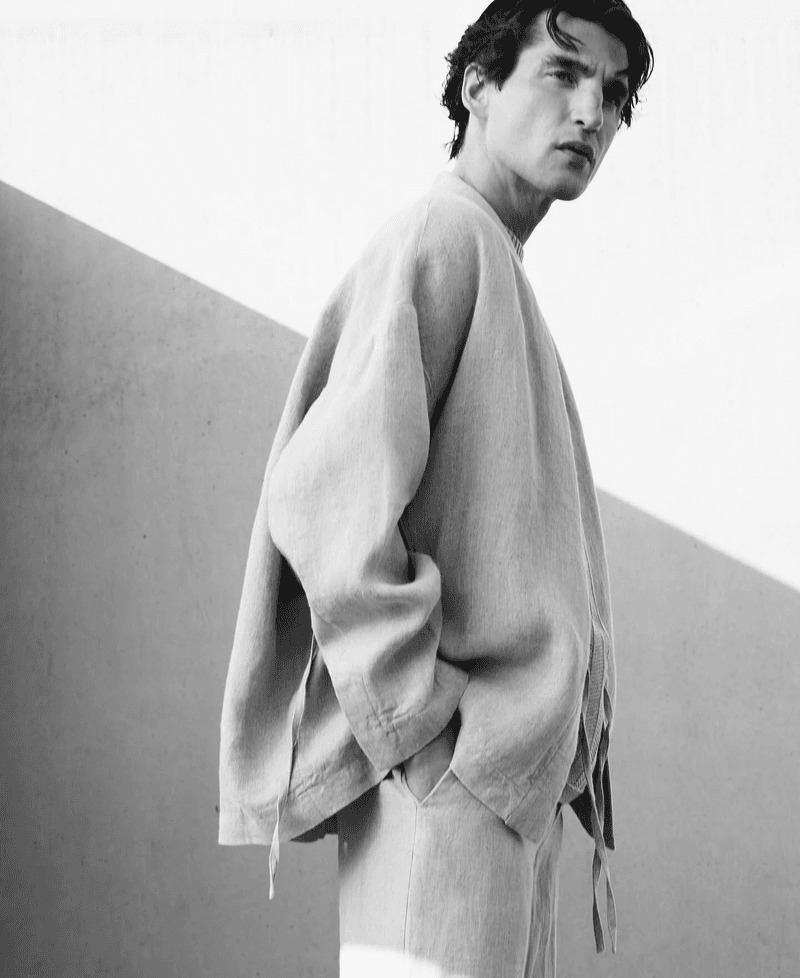 Anatol stands in profile in an oversized natural linen Adolfo Dominguez tunic and matching trousers with raw edges and exposed seams, photographed in black and white