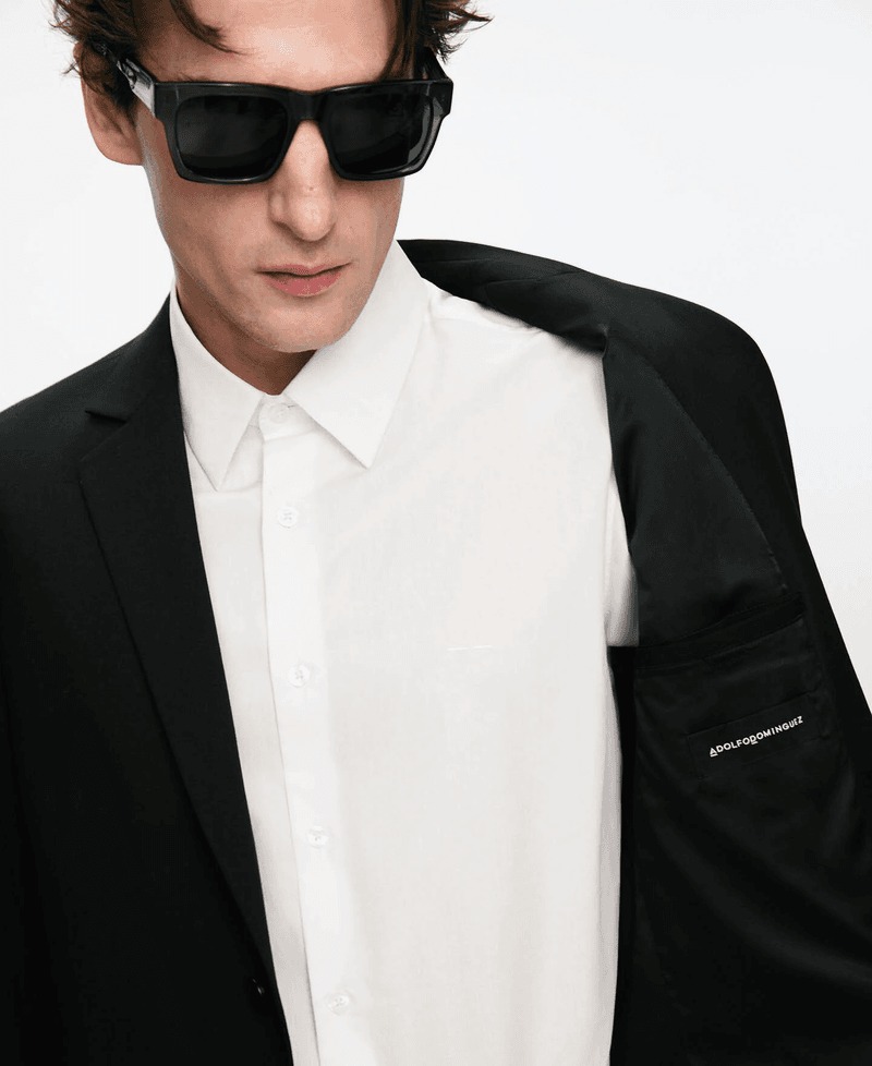 Anatol holds a black blazer over one shoulder, revealing the silk lining and a printed Adolfo Dominguez label, worn over a white shirt with black sunglasses