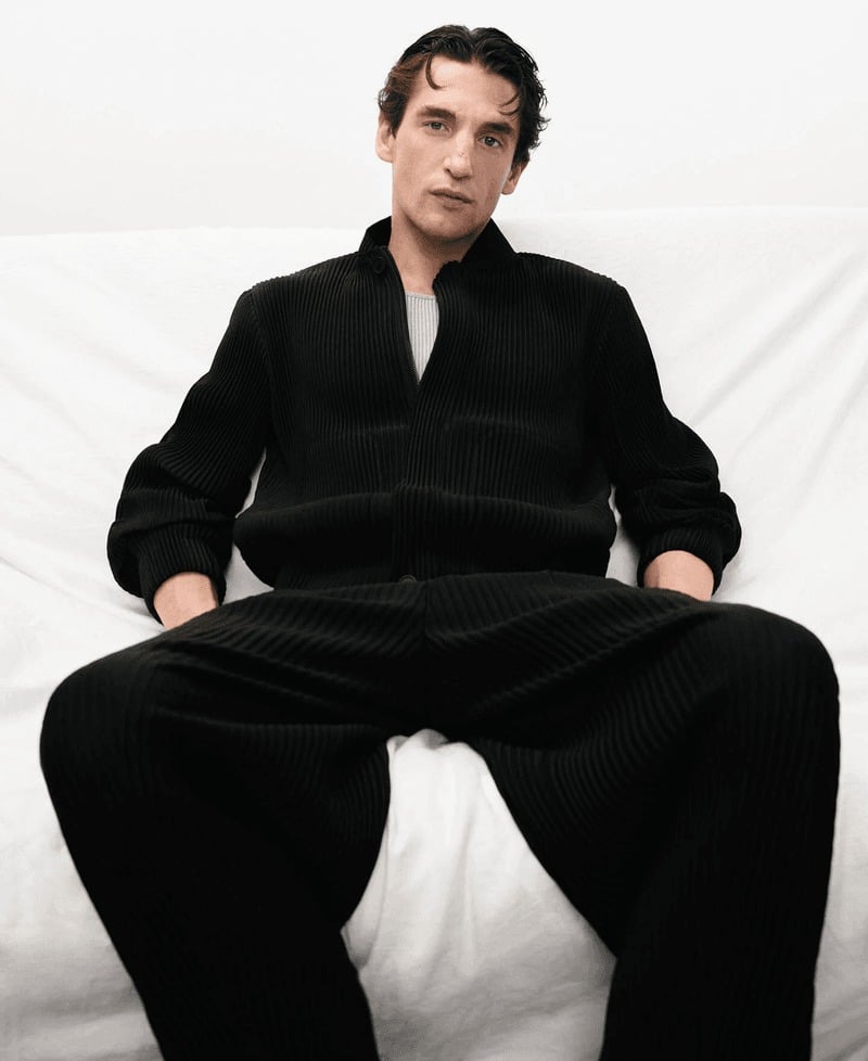 Anatol reclines on a white sofa in a black ribbed-pleat Adolfo Dominguez zip jacket and matching trousers over a gray tee