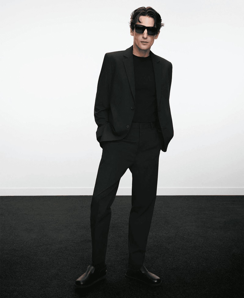Anatol stands in a full-length black Adolfo Dominguez suit with a black t-shirt, black sunglasses, and black leather shoes