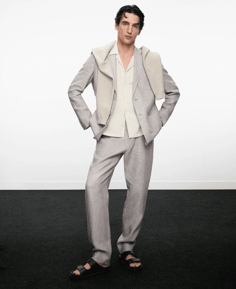 Anatol stands in a light gray linen Adolfo Dominguez suit layered over a cream camp-collar shirt with a hooded scarf and black slide sandals