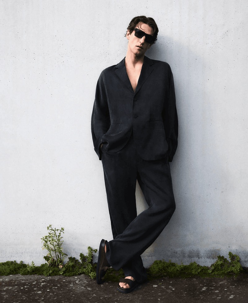 Anatol leans against a weathered wall outdoors in an oversized black linen Adolfo Dominguez suit and black slide sandals with sunglasses