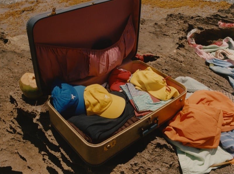 An open vintage tan suitcase rests on rocky sand, packed with folded clothes and capped with a blue baseball hat and a yellow baseball hat, a green coconut and an orange garment scattered alongside it