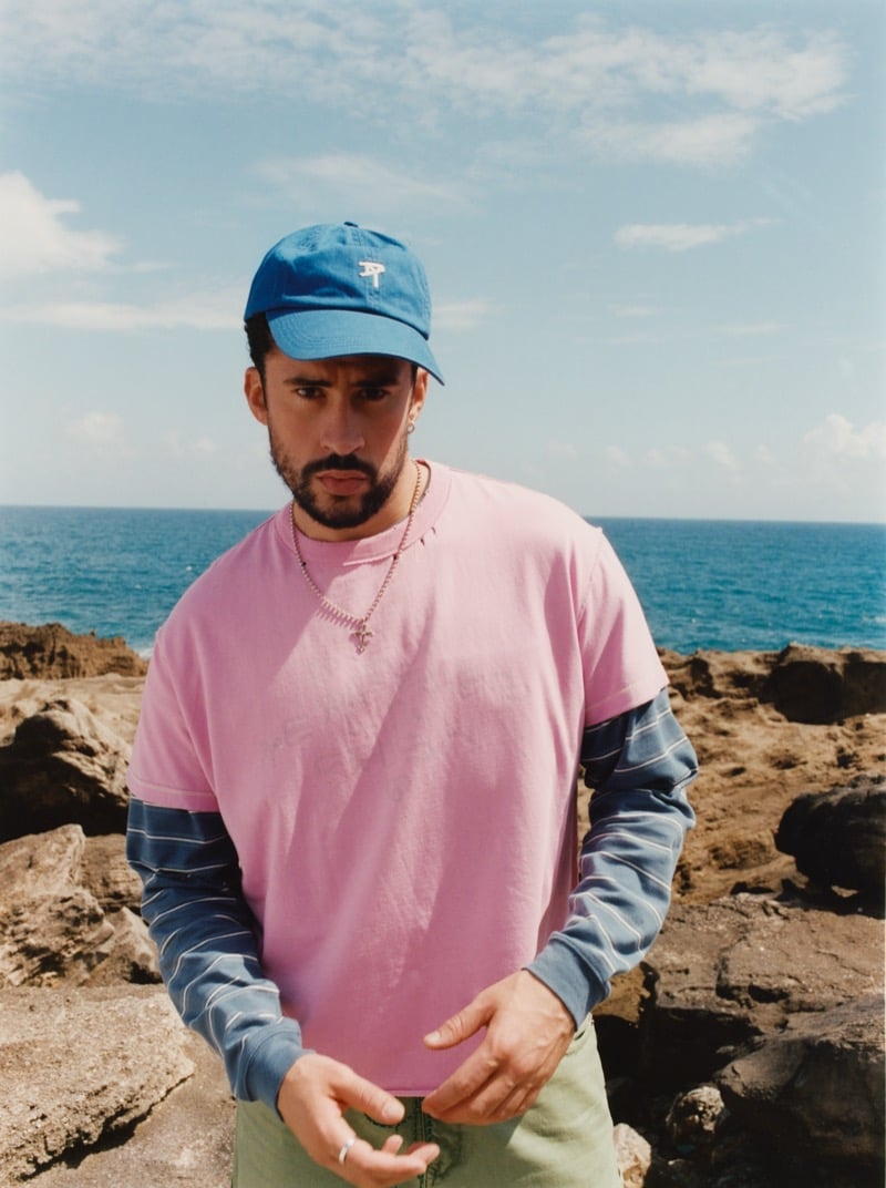 Bad Bunny stands on a rocky Puerto Rican shoreline in a washed blue baseball cap, oversized pink crewneck tee layered over a blue-and-white striped long-sleeve, with a silver chain at his neck
