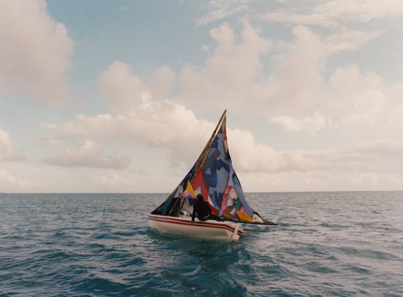 A small handmade wooden sailboat with a patchwork sail glides across the open ocean off the Puerto Rican coast under a soft cloud-streaked sky