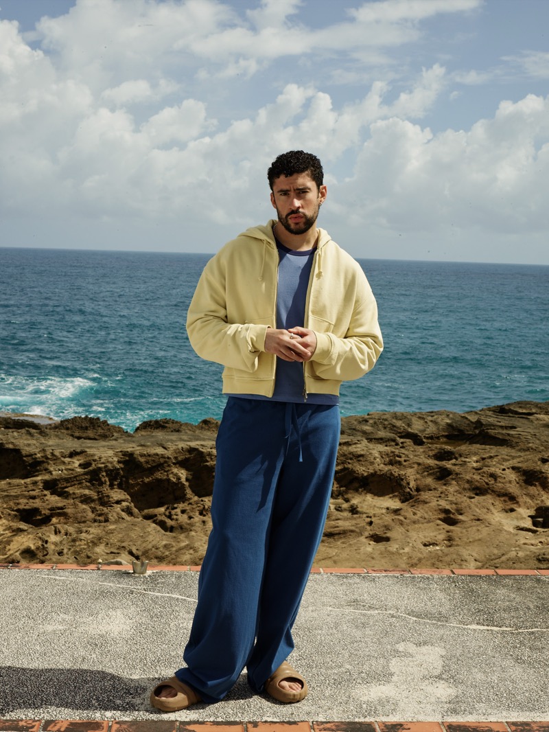 Bad Bunny stands on a coastal terrace in a pale yellow cropped zip-up hoodie open over a slate-blue tee, navy wide-leg drawstring pants, and tan sandals, the ocean and rocky cliffs behind him