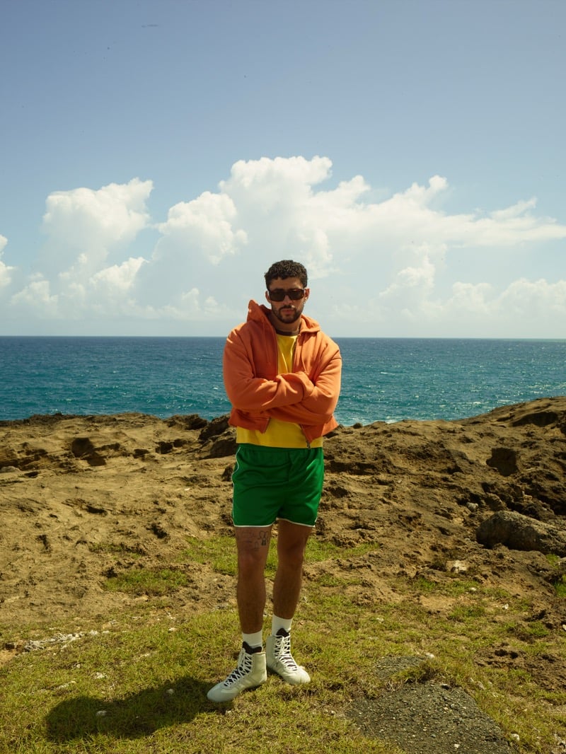 Bad Bunny stands on a grassy clifftop in dark sunglasses with arms crossed, wearing an orange hoodie over a yellow tee, green athletic shorts with a white side stripe, white tube socks, and white high-top sneakers