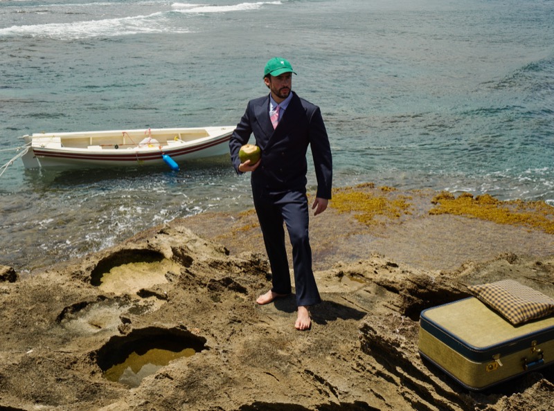 Bad Bunny stands barefoot on a rocky shoreline in a navy double-breasted suit, blue shirt and pink tie, and green baseball cap, holding a green coconut, a small wooden boat moored behind him and a vintage suitcase set on the rocks to his right