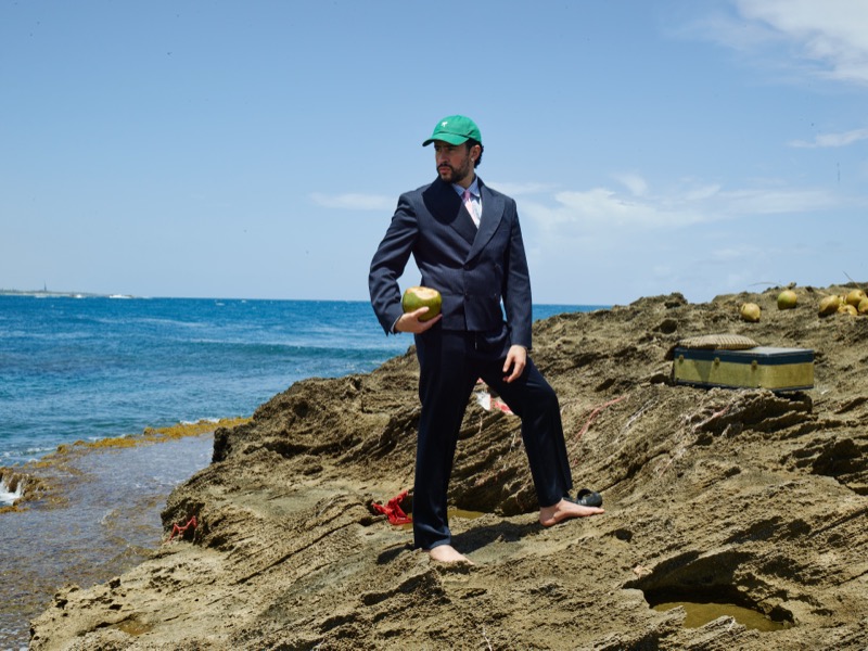 Bad Bunny walks barefoot across a sandy rocky outcrop above the ocean in a navy double-breasted suit and green baseball cap, holding a coconut, a vintage olive trunk and scattered coconuts in the background