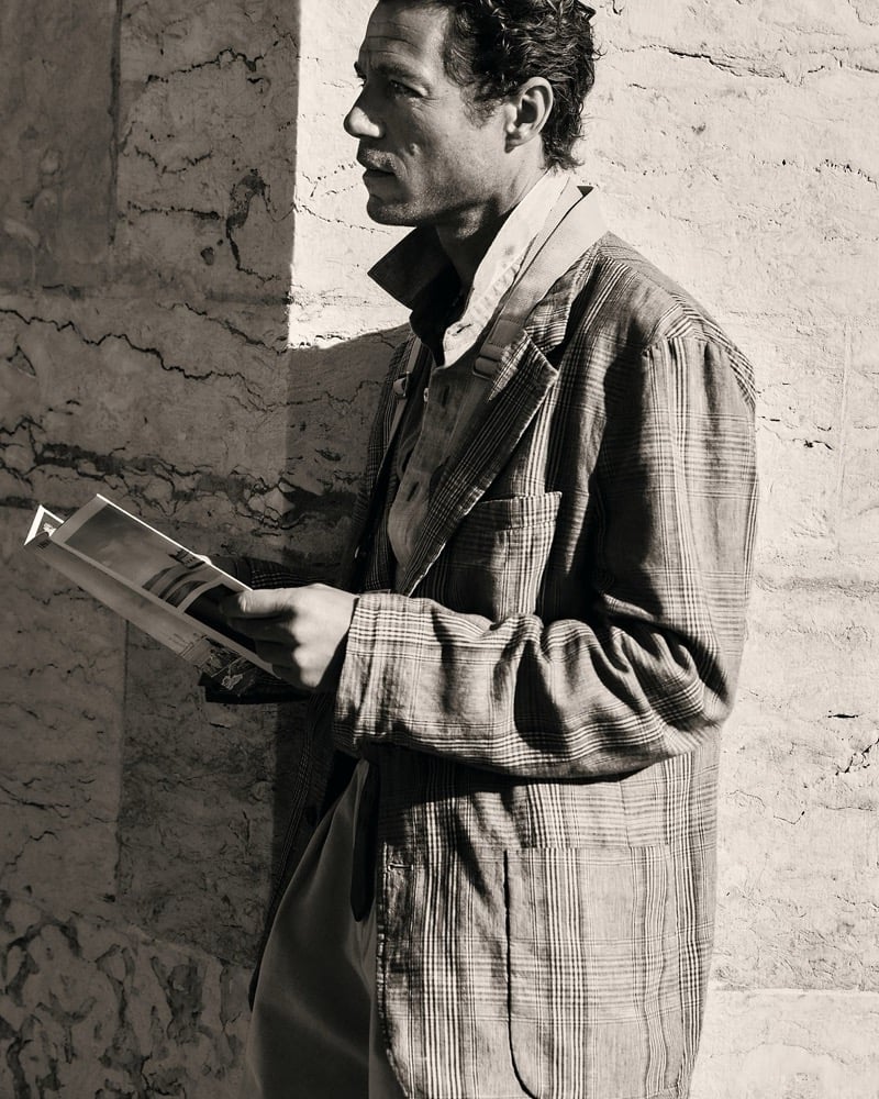Sepia profile of Jon Paul leaning against a weathered stone wall reading a magazine in a windowpane plaid linen jacket over an open collared shirt with relaxed trousers