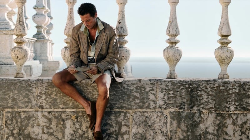 Jon Paul sits on a stone balustrade overlooking the sea reading a brochure in a brown plaid linen blazer over a color block linen shirt with cream shorts, a Leica around his neck, and brown suede loafers