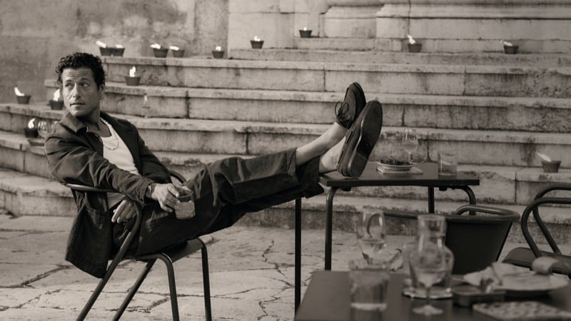 Sepia image of Jon Paul reclining at an outdoor Lisbon cafe with his feet propped on a chair in a dark linen jacket and dark trousers with leather loafers, holding a drink