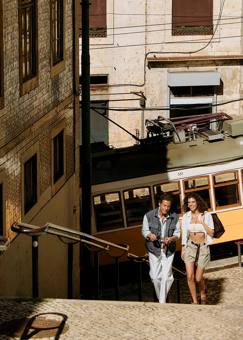 Jon Paul climbs cobbled Lisbon steps with a female model as a yellow Lisbon tram passes behind them, wearing a slate gray quilted vest over a chambray shirt with white linen trousers